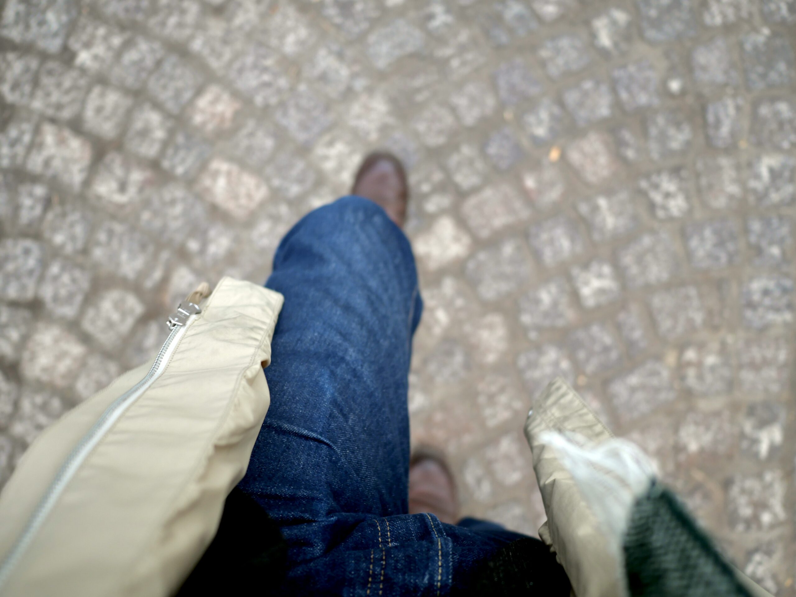 A photograph from just above my waist looking down onto a cobblestone path taken mid-step. You can see my blue jeans, brown boots, and parts of my rain coat and scarf.